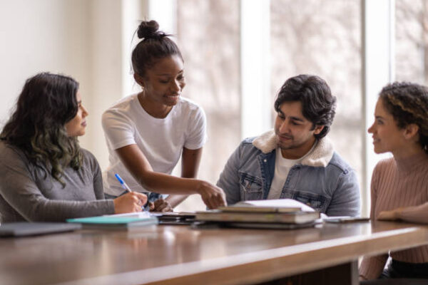 A young female University student of African decent, stands behind a peer and leans in as she tries to help her classmate with his studies. They are both dressed casually and are among a group sitting at the table all studying together for class.